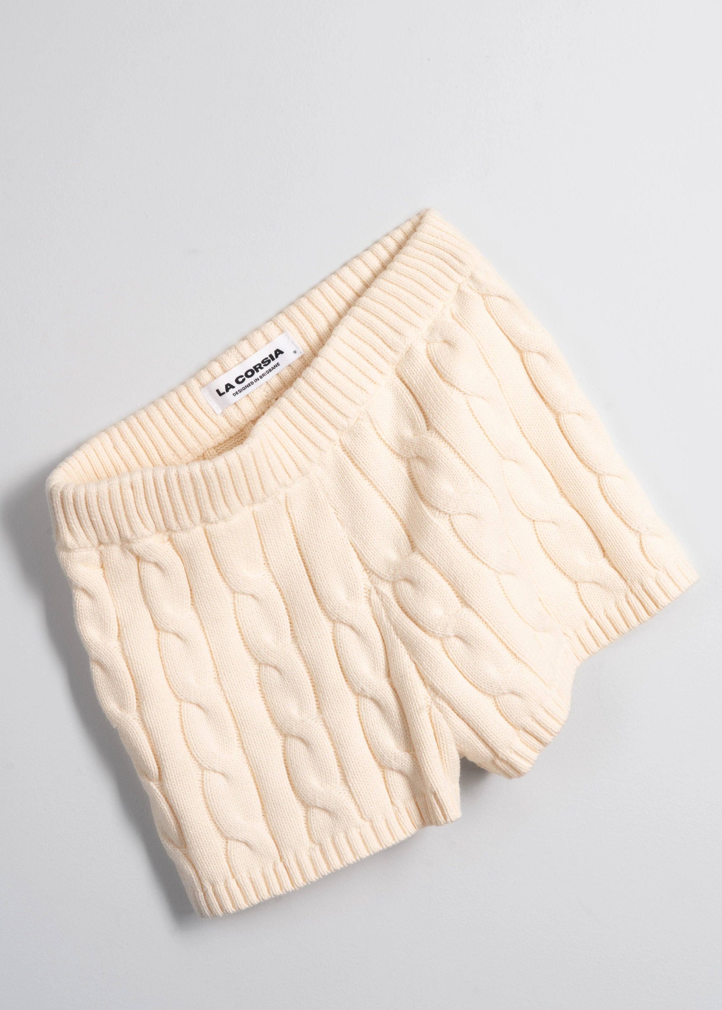 La Corsia Cable Knit Mini Short in cream, made from heavyweight cotton with a fitted silhouette, ribbed waistband, and chunky cable texture throughout.
