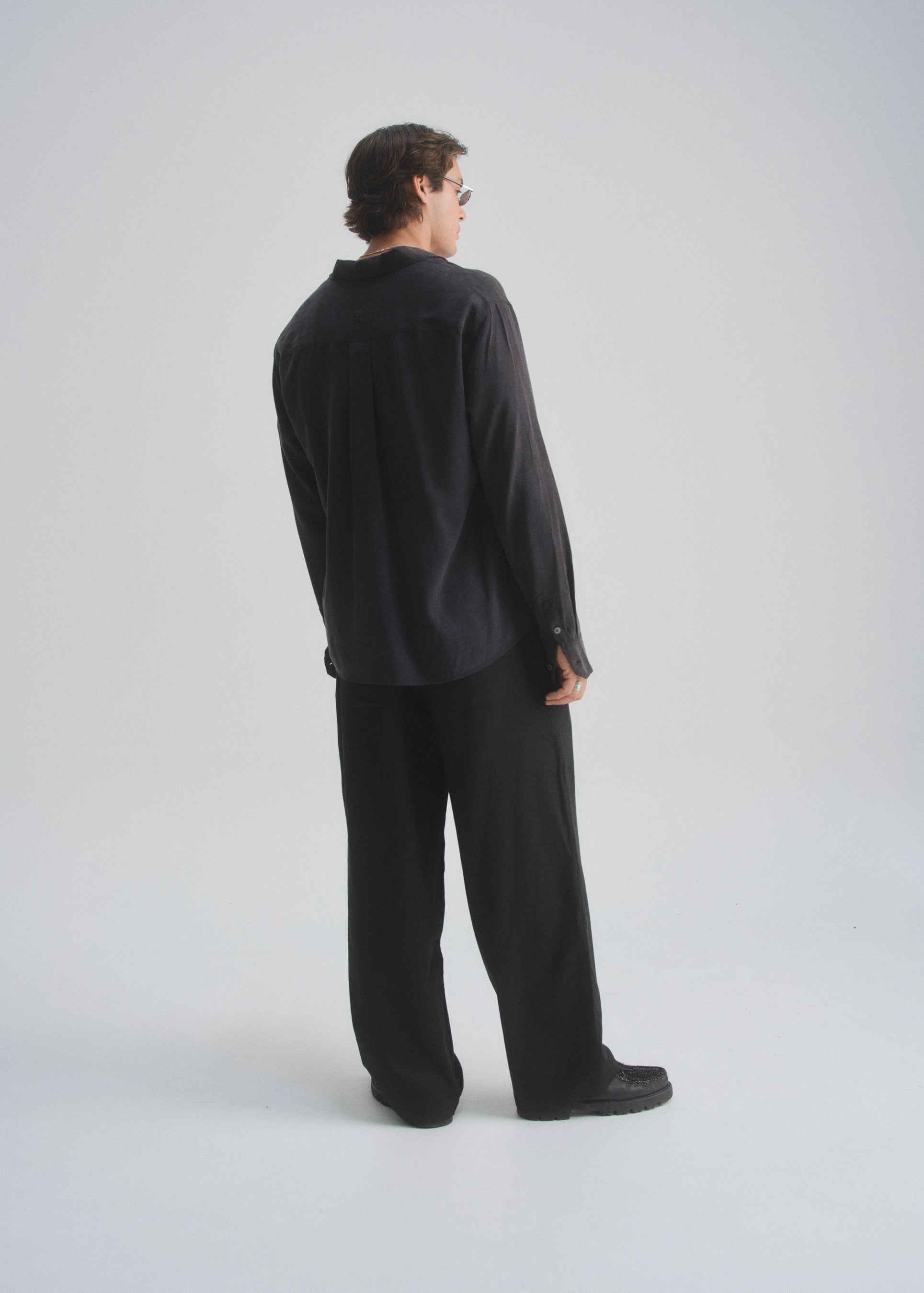 Man wearing a black button up shirt and wide leg pants standing against a white background