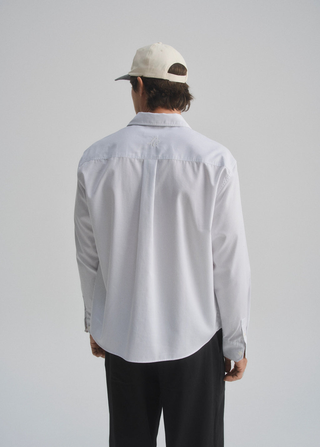 Person wearing a pinstripe white and navy shirt and dark pants with a white cap on a plain background