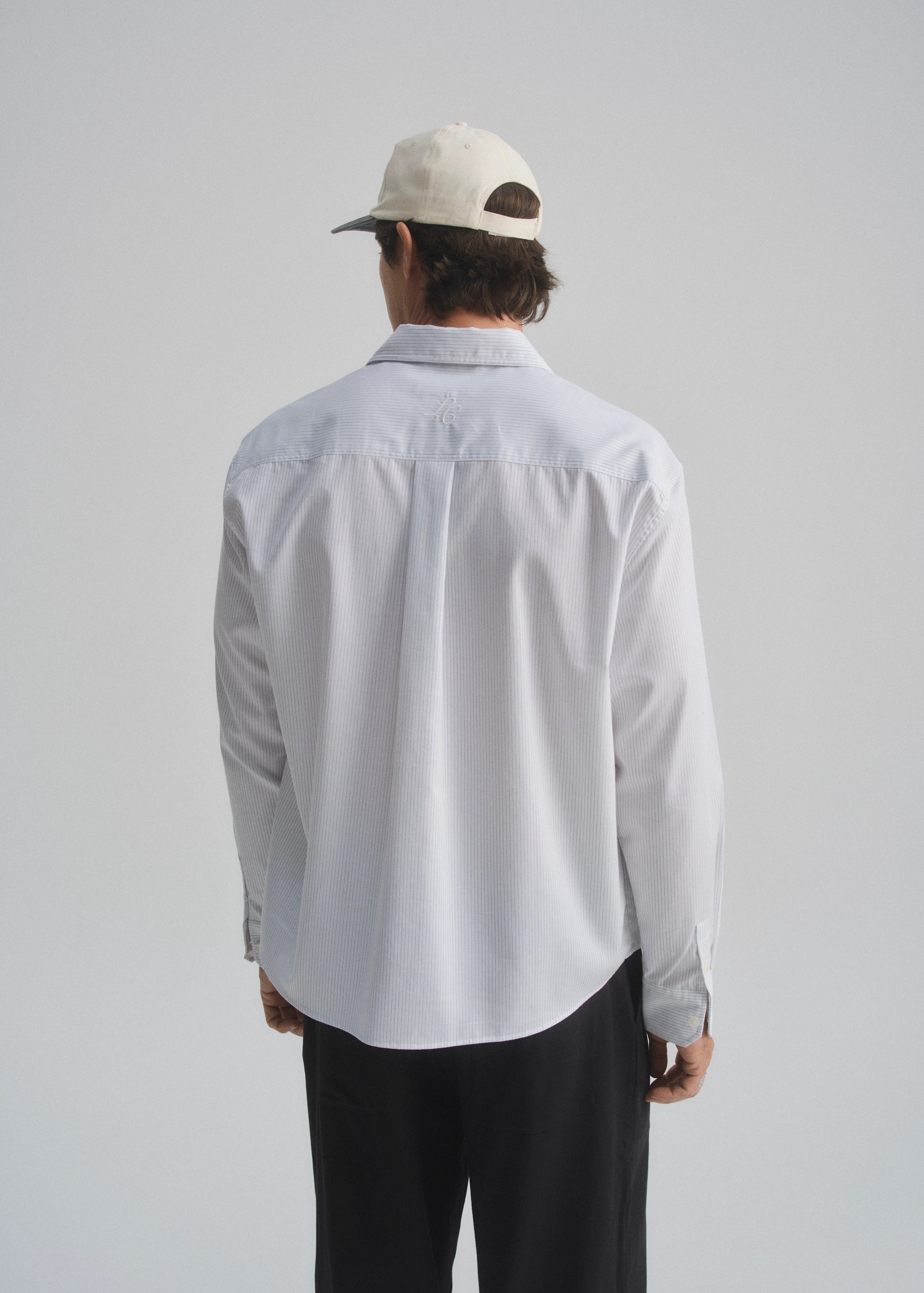 Person wearing a pinstripe white and navy shirt and dark pants with a white cap on a plain background