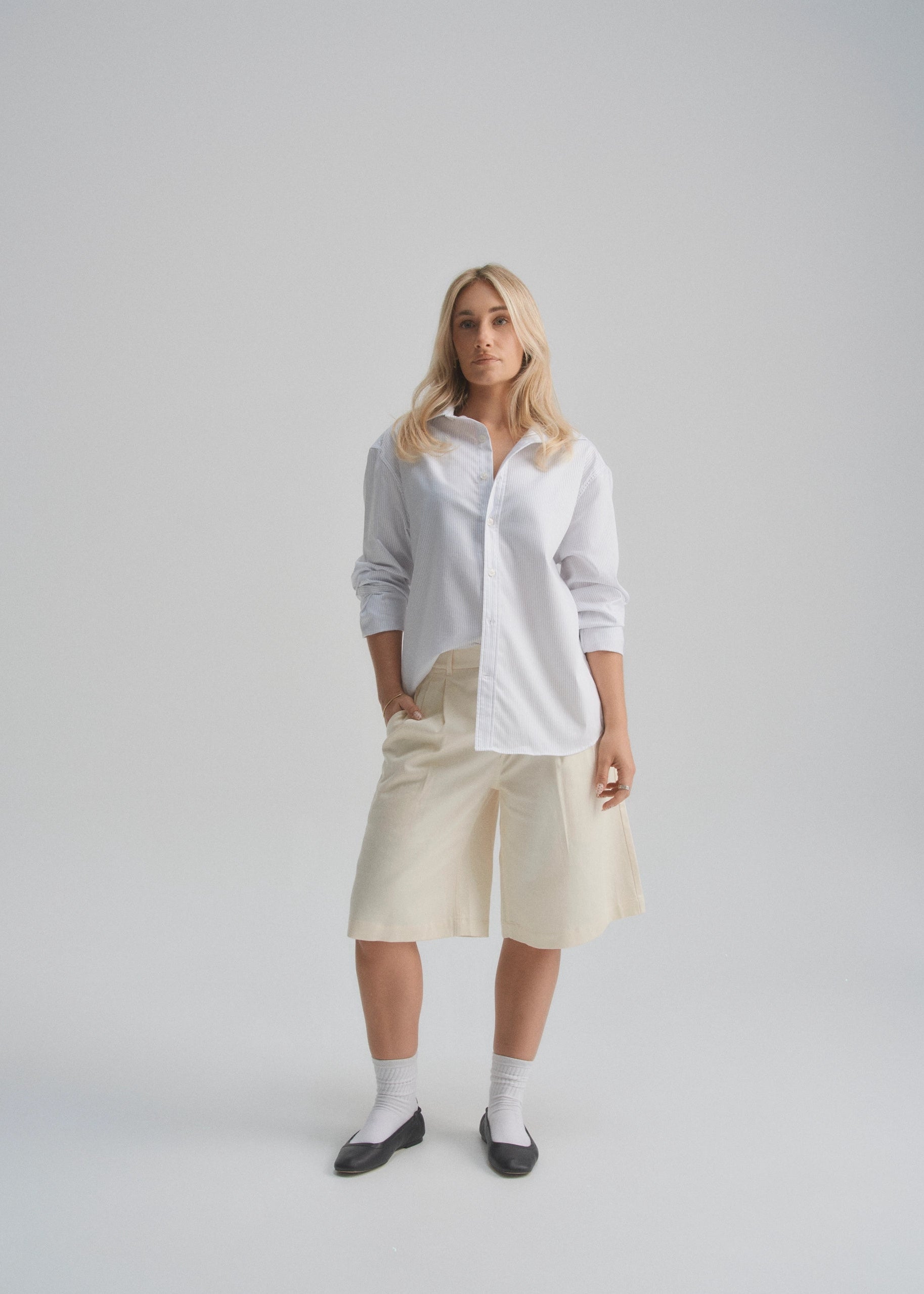 Woman wearing a white shirt and beige wide leg pleated shorts on a plain background