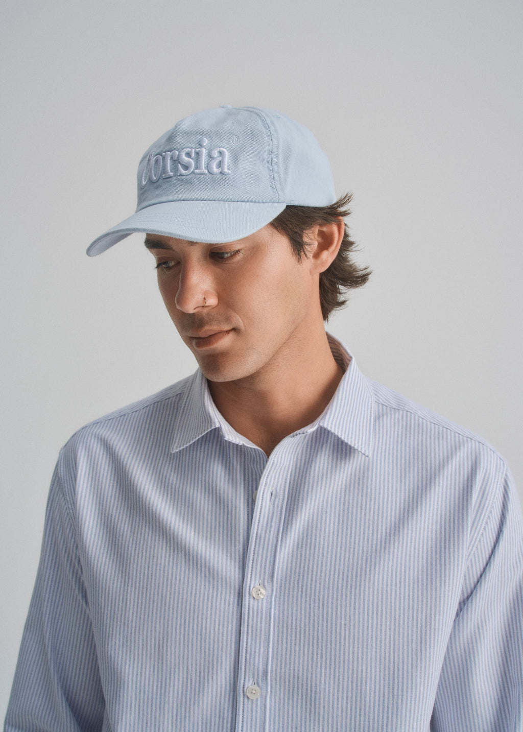 Man wearing a light blue cap with 'Corsia' branding and a light blue striped shirt on a gray background