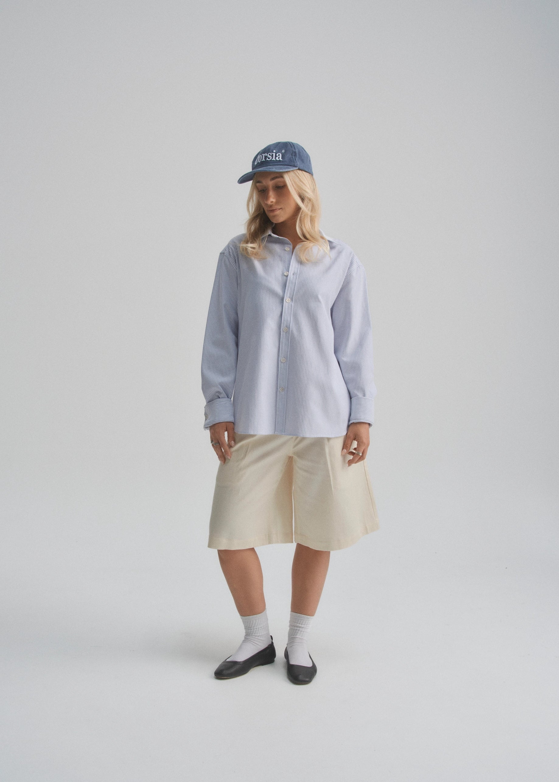 Model wearing a light blue shirt, wide leg beige pleated shorts, and black shoes on a white background