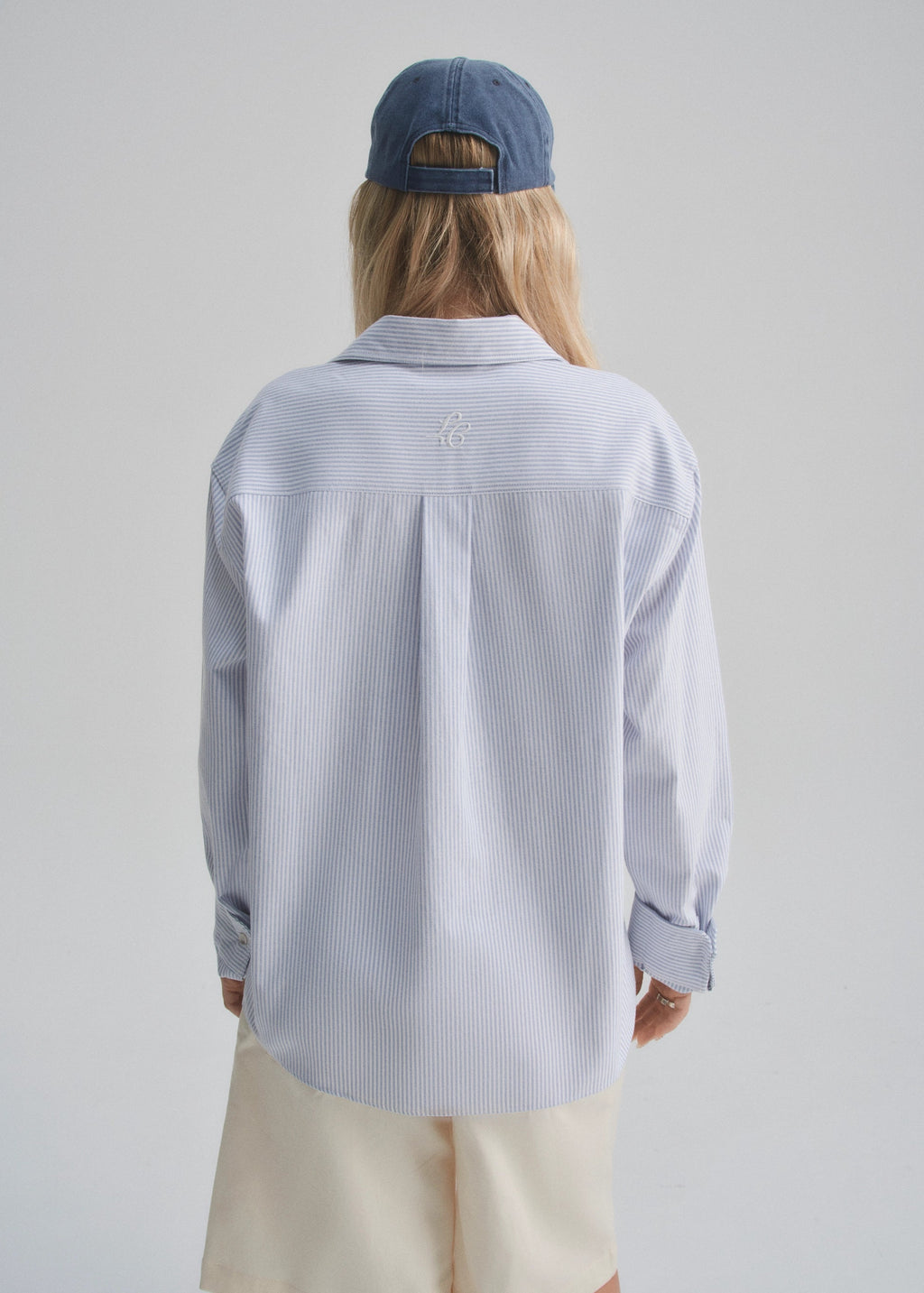 Person wearing a light blue striped shirt and beige pants with a faded navy cap on a plain background