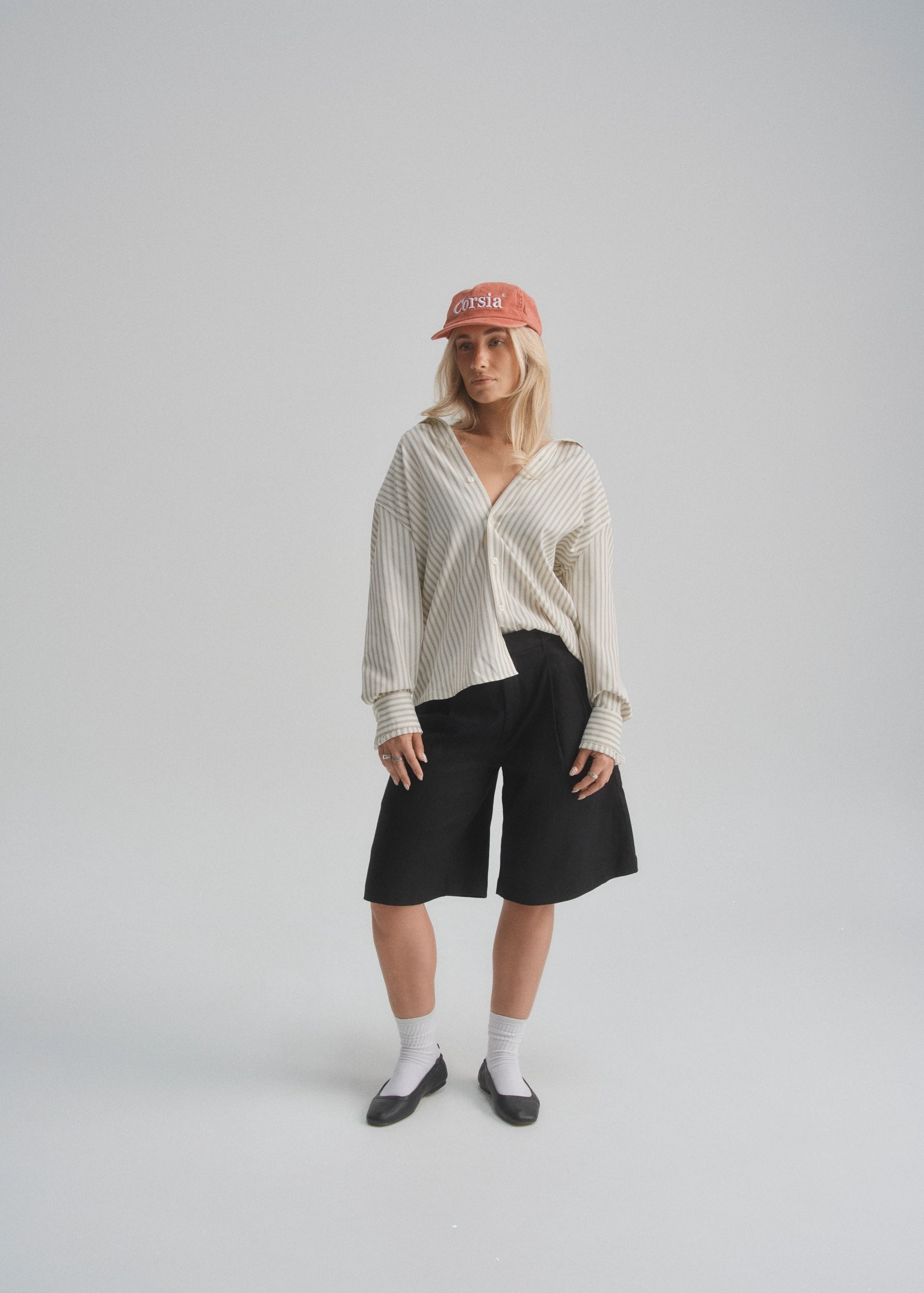 Person wearing a cream and brown striped shirt, black shorts, and a red cap on a plain background