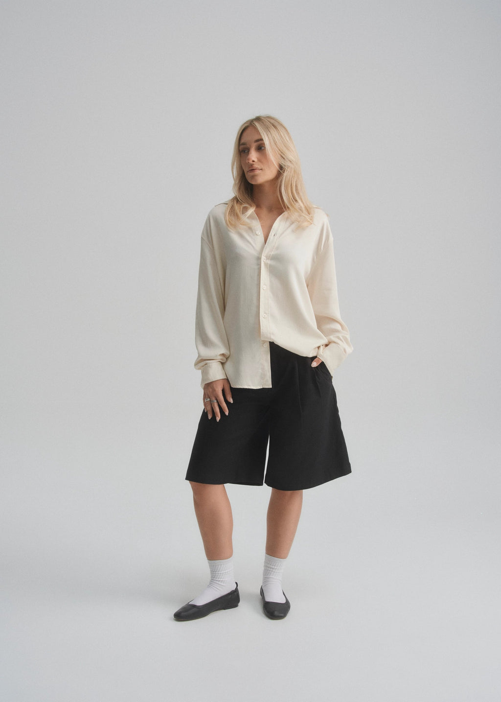 Woman wearing a cream blouse, black wide leg shorts, and black leather shoes on a plain background