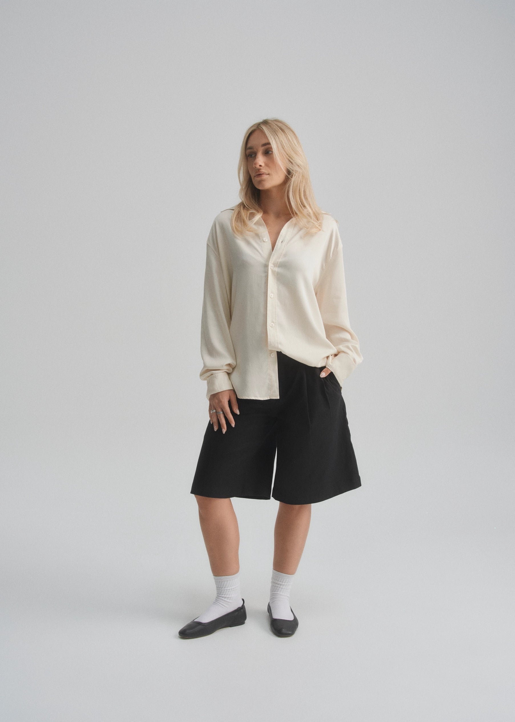 Woman wearing a cream blouse, black wide leg shorts, and black leather shoes on a plain background