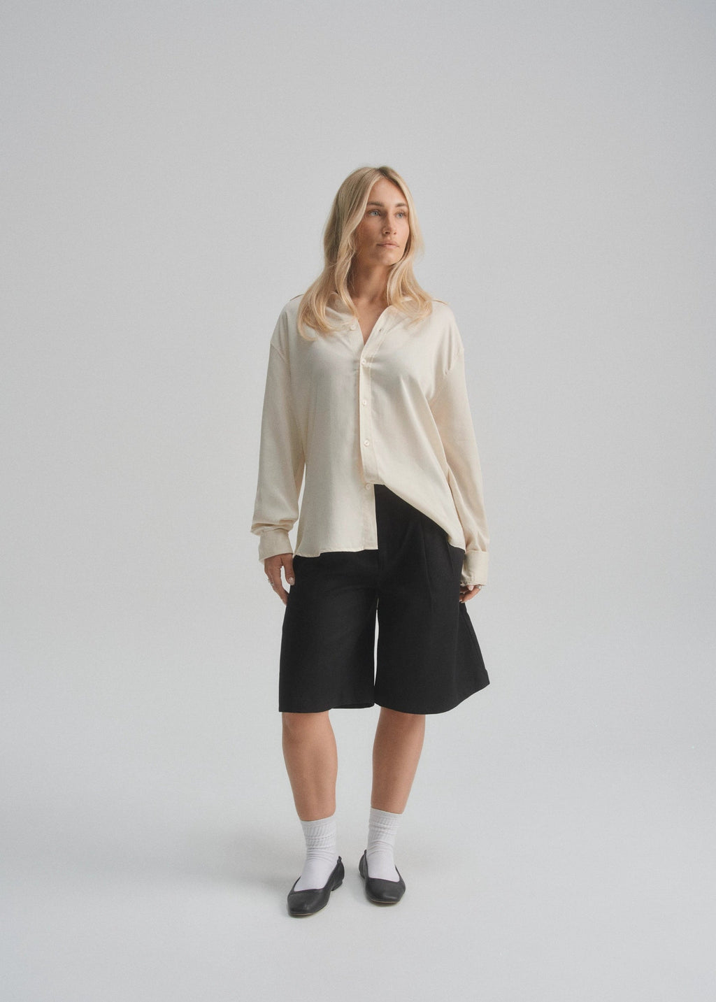 Woman wearing a cream button up shirt and black shorts on a plain background