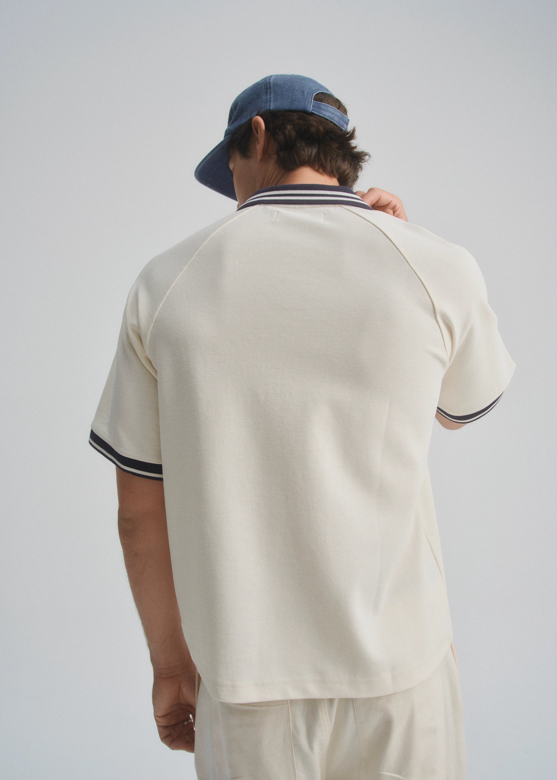 Person wearing a beige polo shirt with navy trim and a blue cap on a plain background