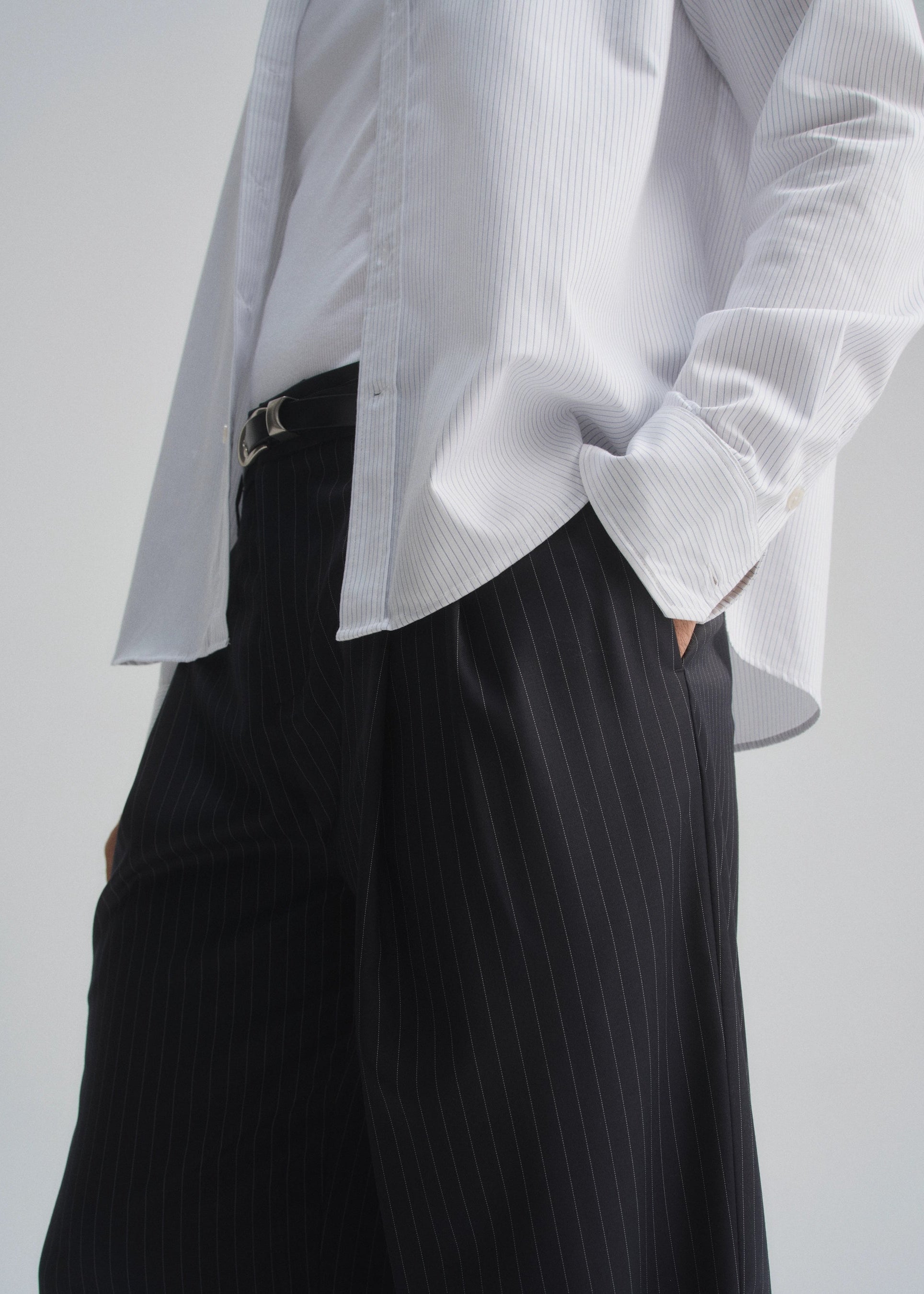 Person wearing a white pinstripe shirt and dark navy pleated pants on a plain background