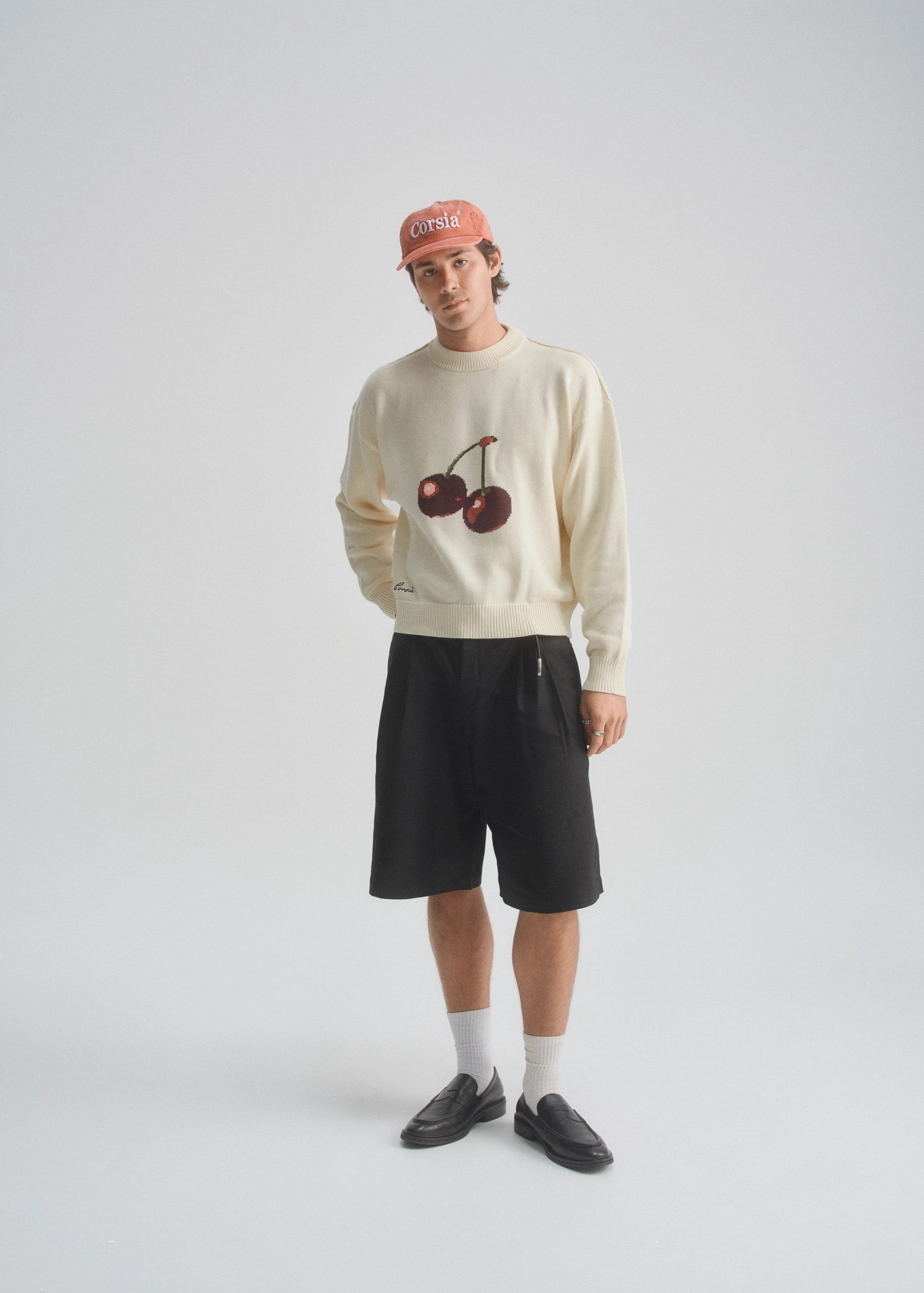 Person wearing a cream sweater with cherry design, black shorts, and a pink cap on a white background