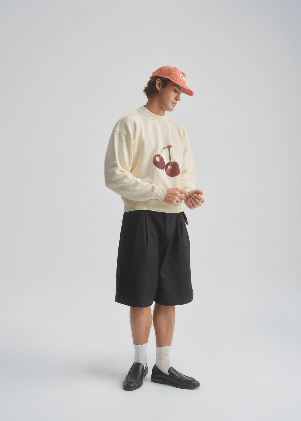 Person wearing a cream sweatshirt with a cherry graphic design, black shorts, and a red cap on a white background