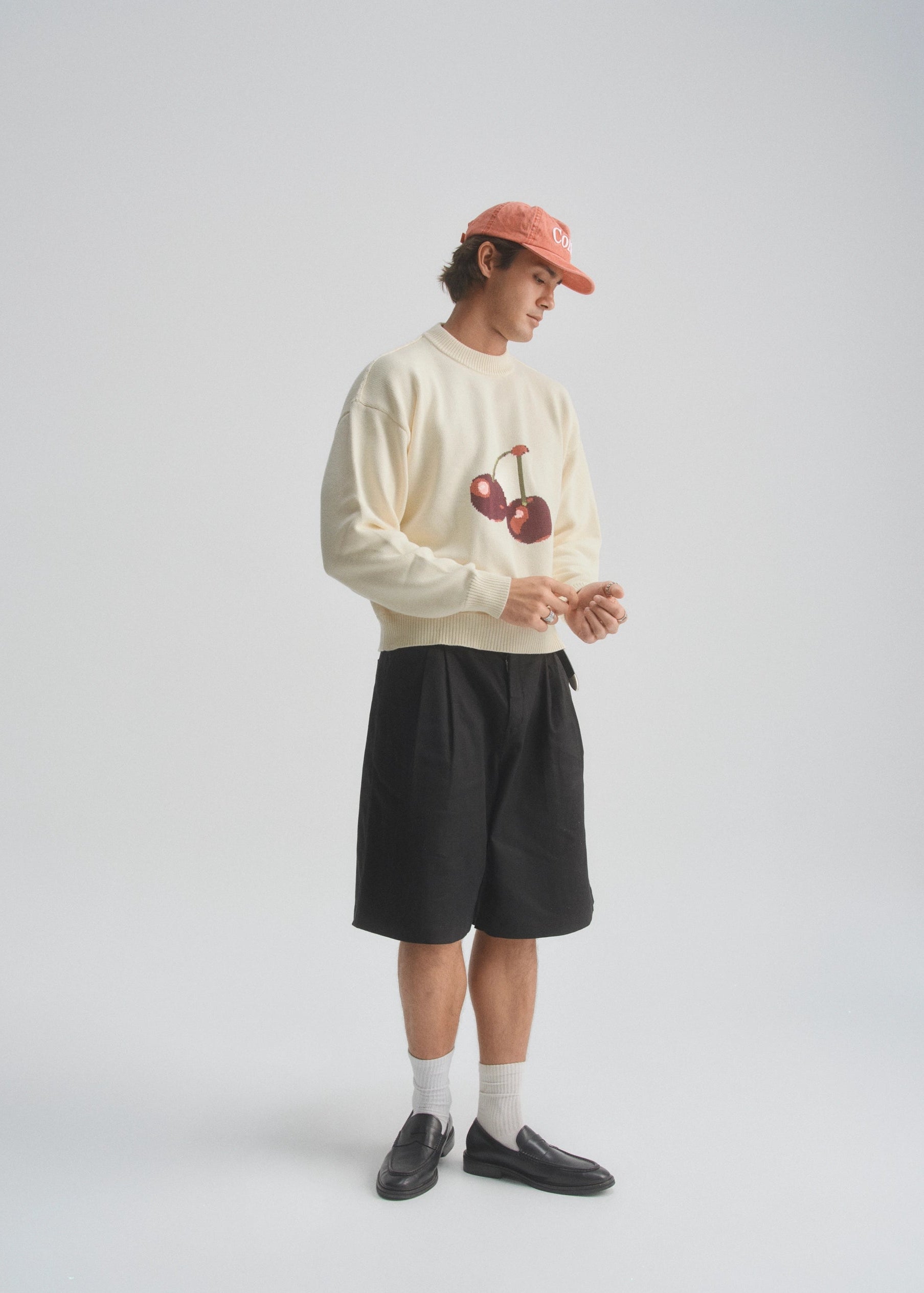 Person wearing a cream sweatshirt with a cherry graphic design, black shorts, and a red cap on a white background