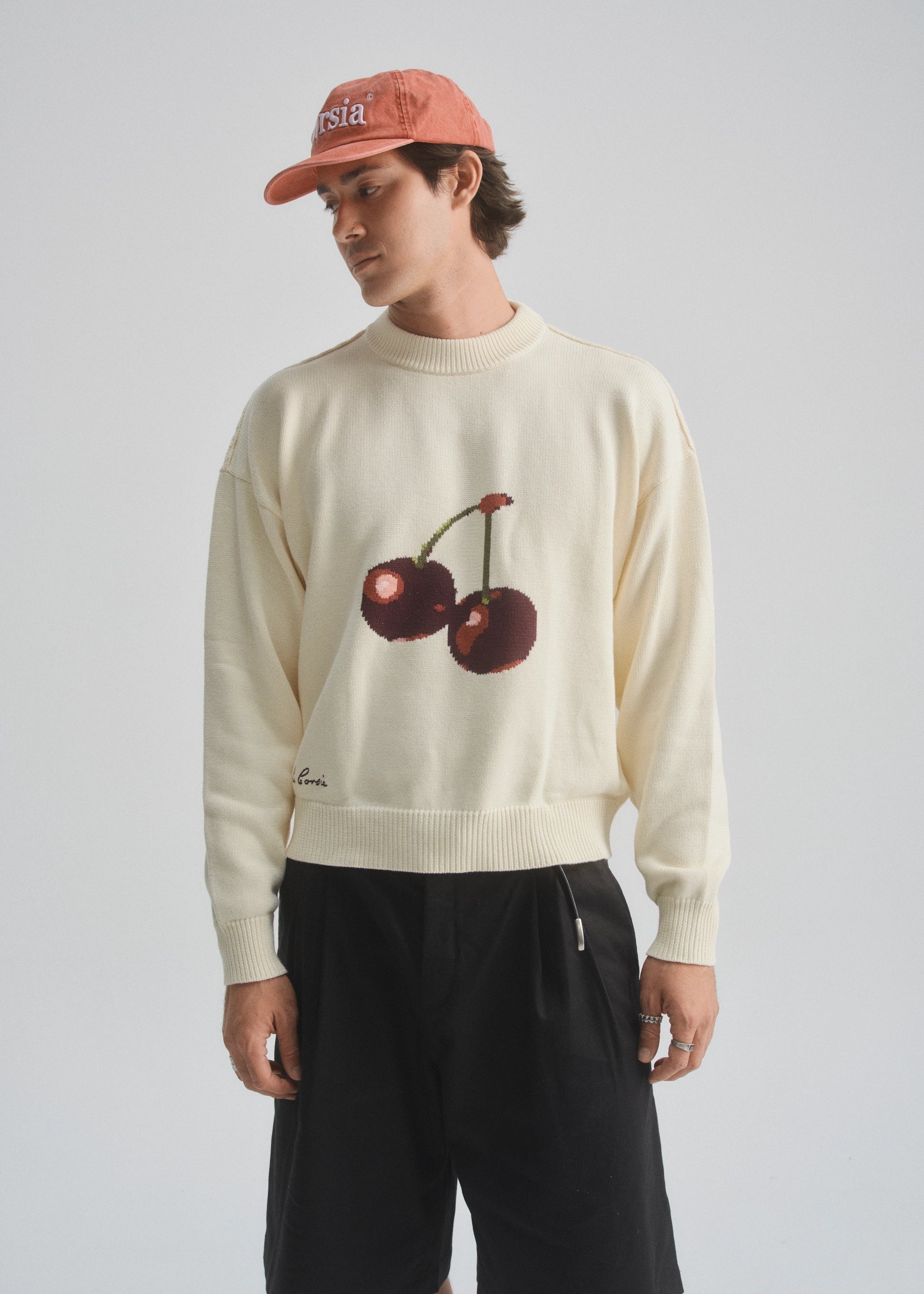 Person wearing a cream sweater with cherry design and a red cap on a light gray background