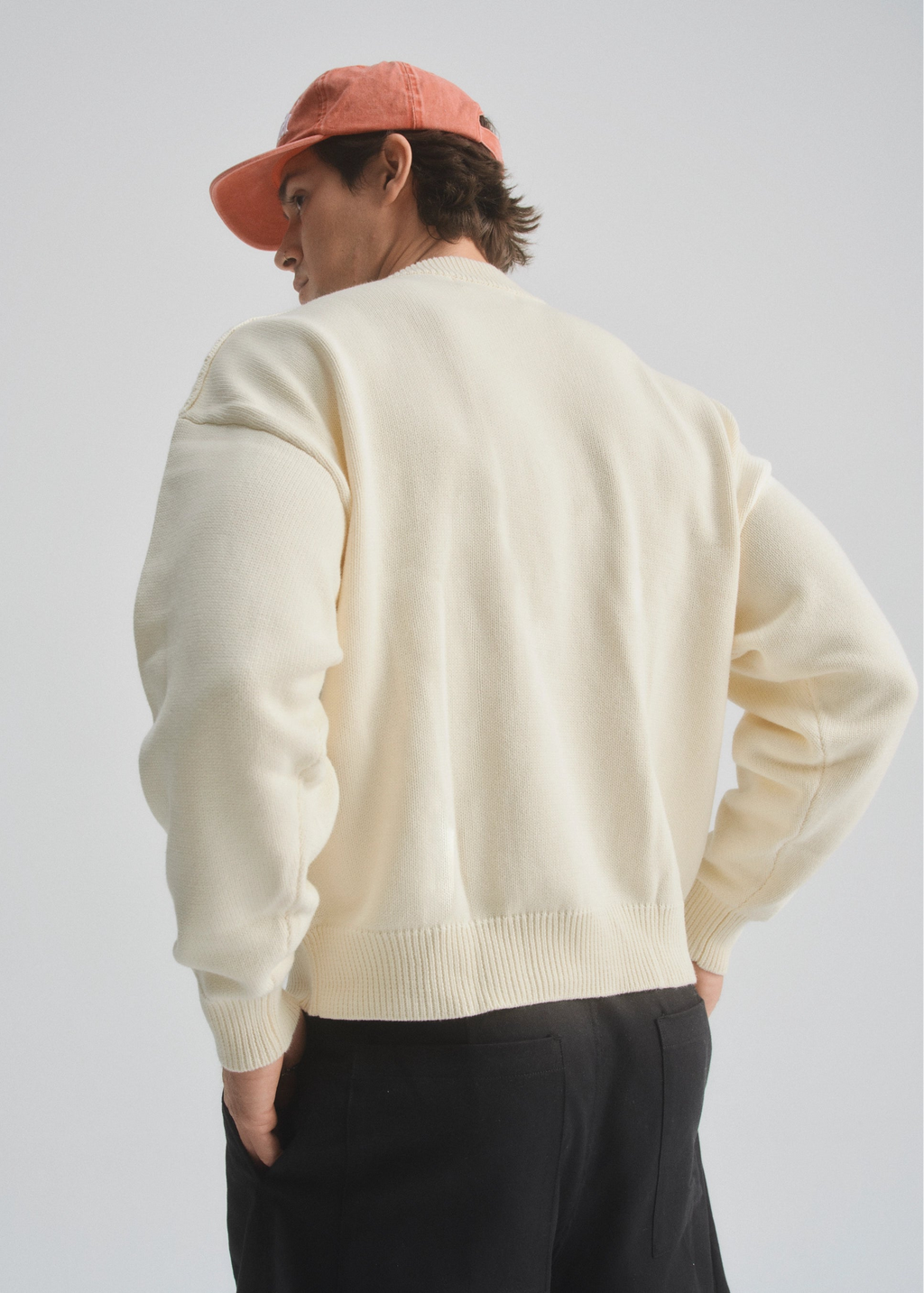 Person wearing a cream sweater and faded red cap, and black shorts on a plain background