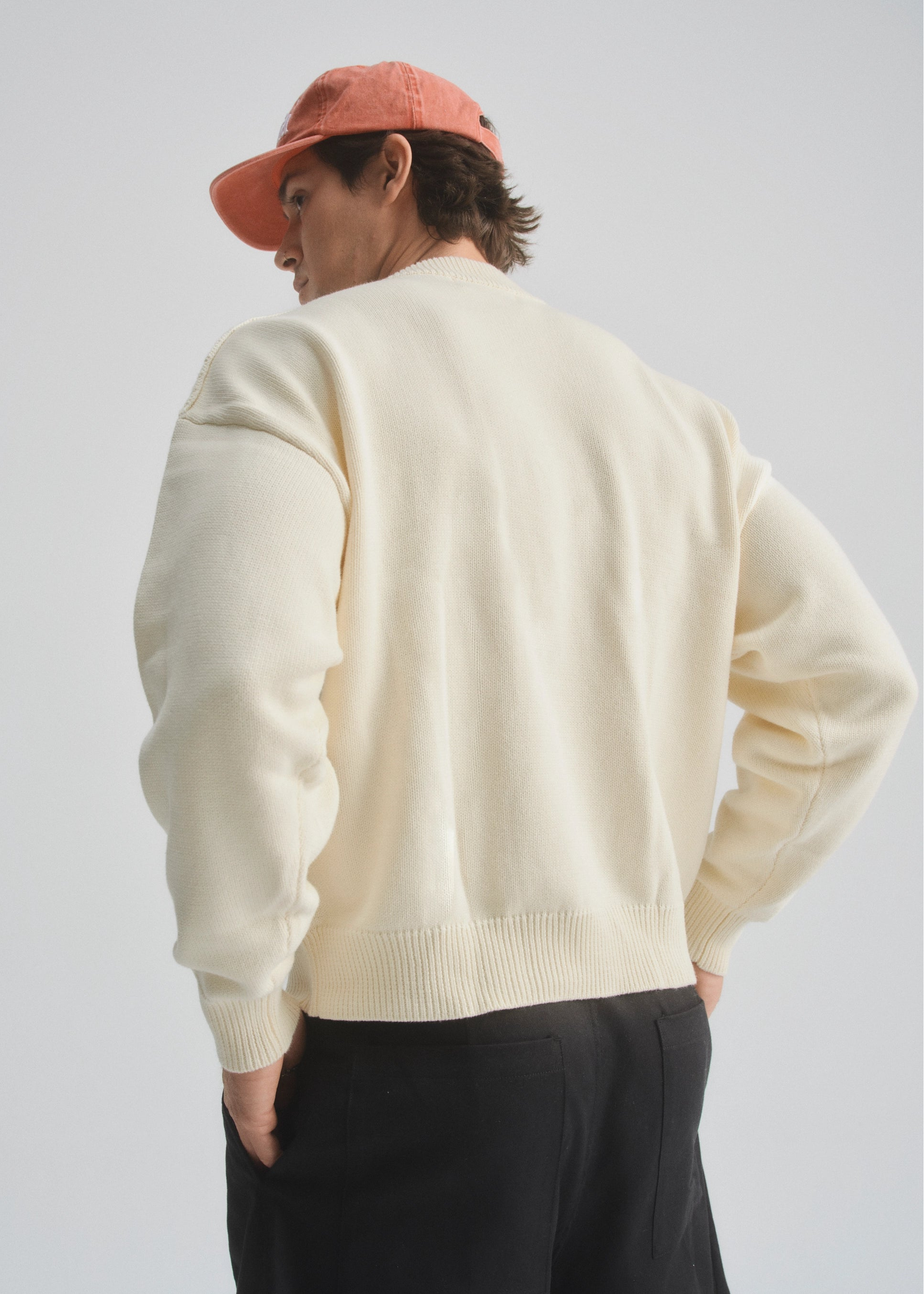 Person wearing a cream sweater and faded red cap, and black shorts on a plain background