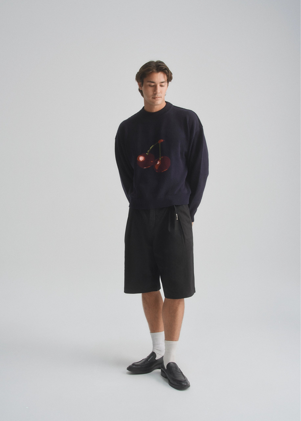 Person wearing a dark navy sweater with a cherry design and black shorts on a white background