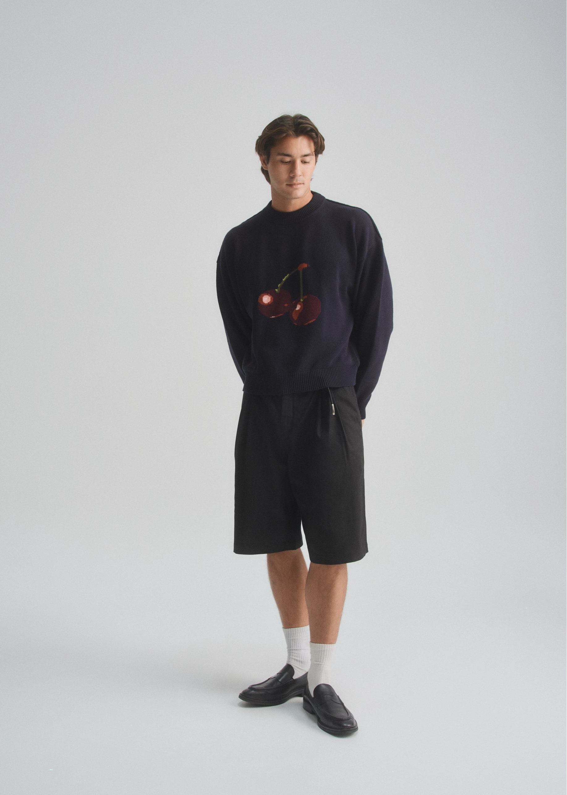 Person wearing a dark navy sweater with a cherry design and black shorts on a white background