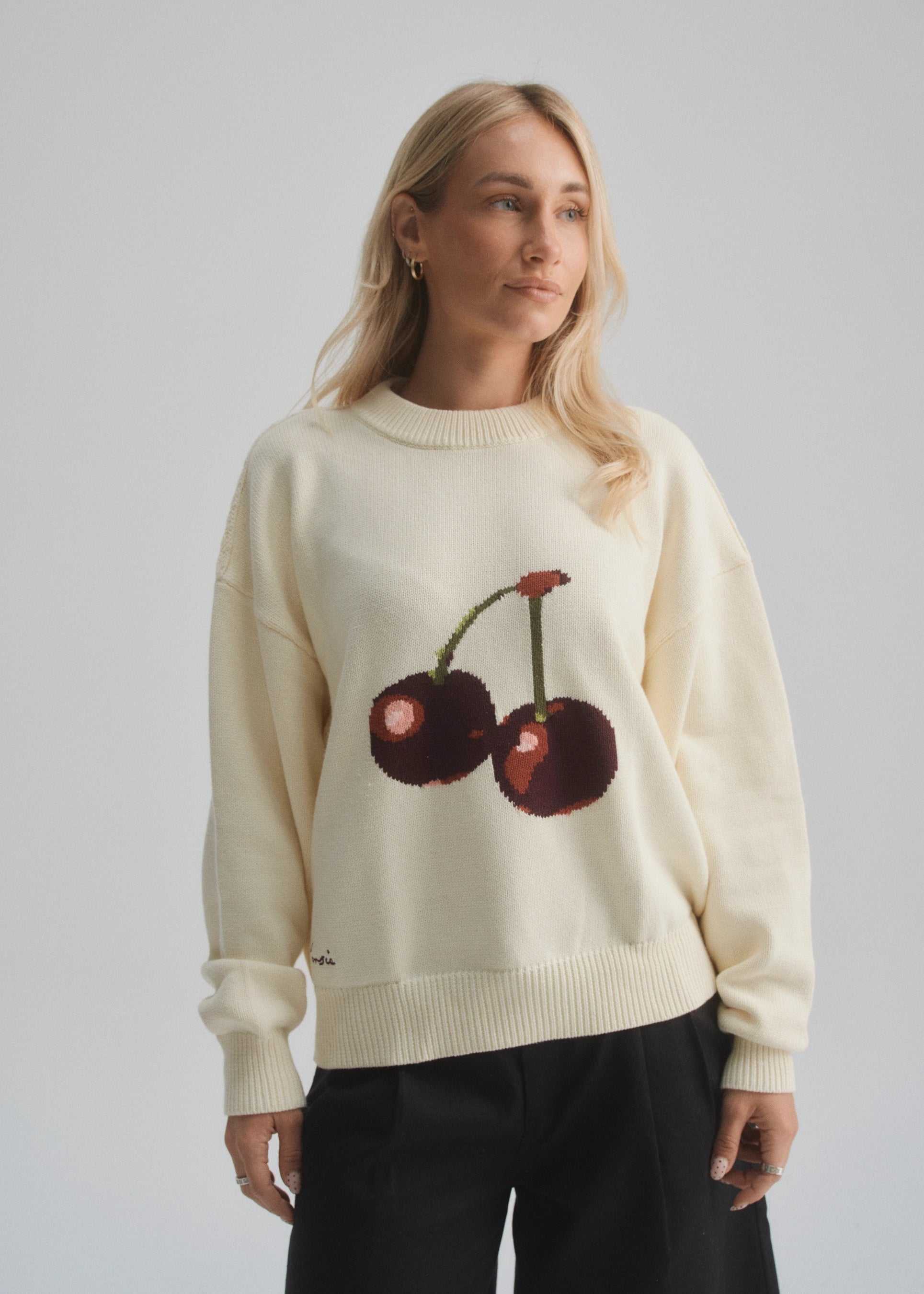 Woman wearing a cream sweater with cherry design against a plain background