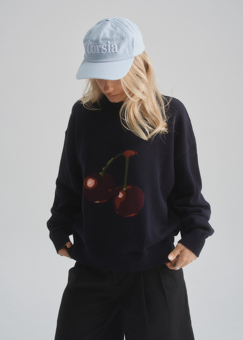Person wearing a black sweater with cherry design and light blue cap on a plain background