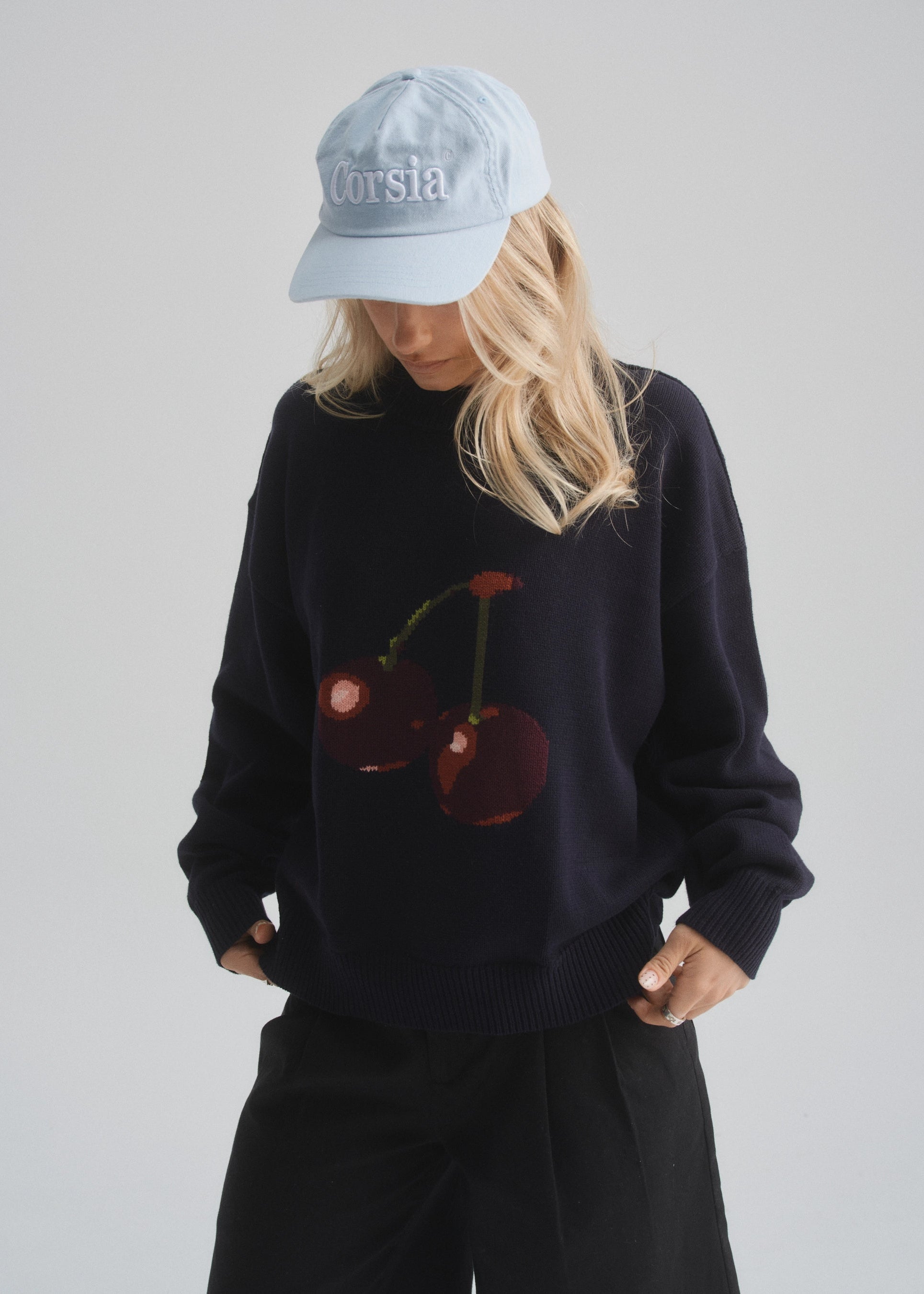 Person wearing a black sweater with cherry design and light blue cap on a plain background