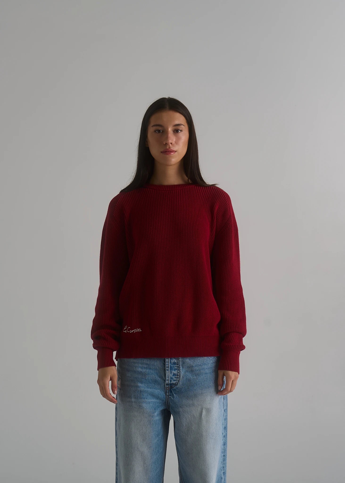 A woman modeling a dark red ribbed knit sweater with a small white embroidered logo on the lower left front, paired with light blue jeans, against a plain light gray background.