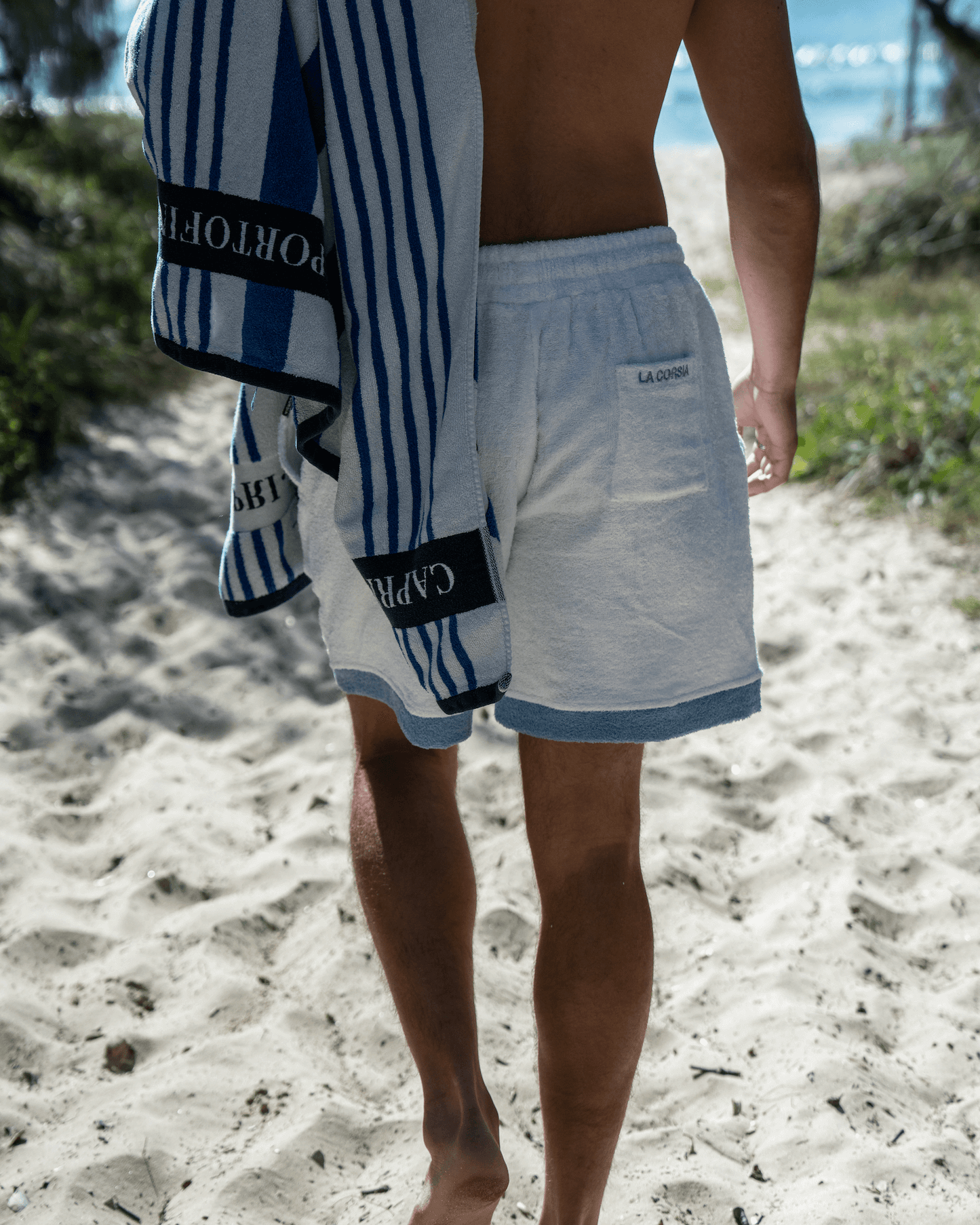 Beach picture on a path, wearing terry towel shorts, towel over shoulder. Loose Fit cotton towelling, embroidery. Custom