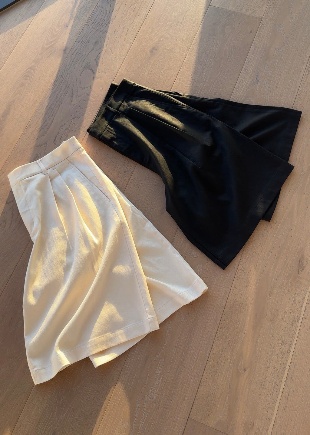 Two shorts folded, one cream and one black, on a wooden floor.